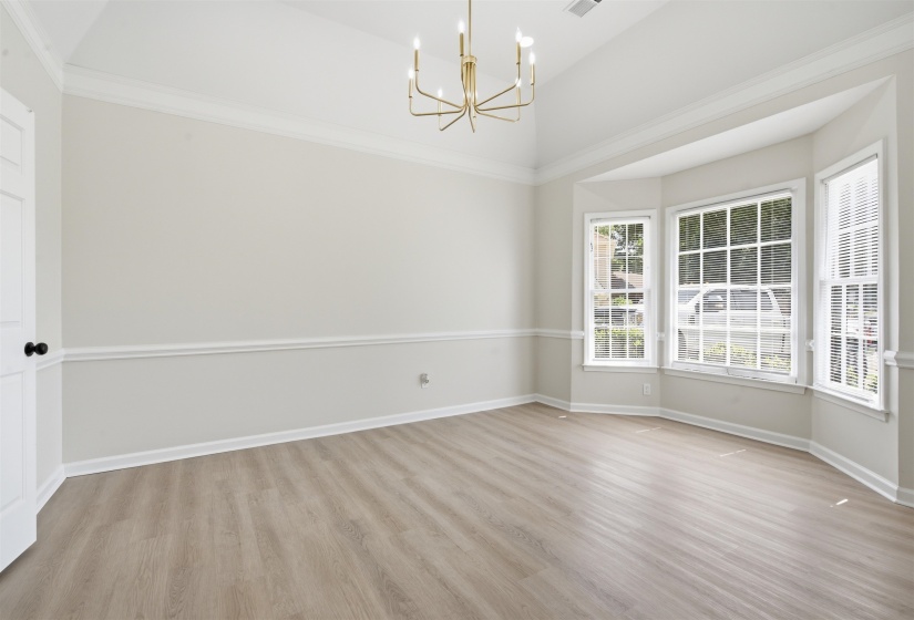 Spacious room featuring a prominent bay window, modern chandelier, vaulted ceiling, wood-finish flooring, and crown molding