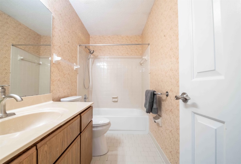 Bathroom featuring a wood-finish vanity with an integrated sink and brushed nickel faucet, a large wall-mounted mirror, a white bathtub with a shower, and tiled flooring