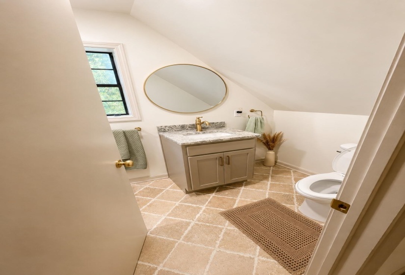 Neutral-toned bathroom with an angled ceiling, featuring a vanity with a gray cabinet and a light-toned countertop, a round gold-framed mirror, and a window with dark trim