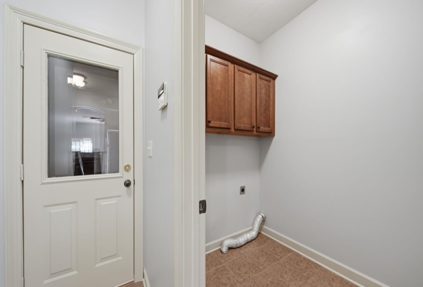 Utility room featuring a white paneled door with a glass insert, light gray walls, warm wood-finish cabinetry, and square floor tiles
