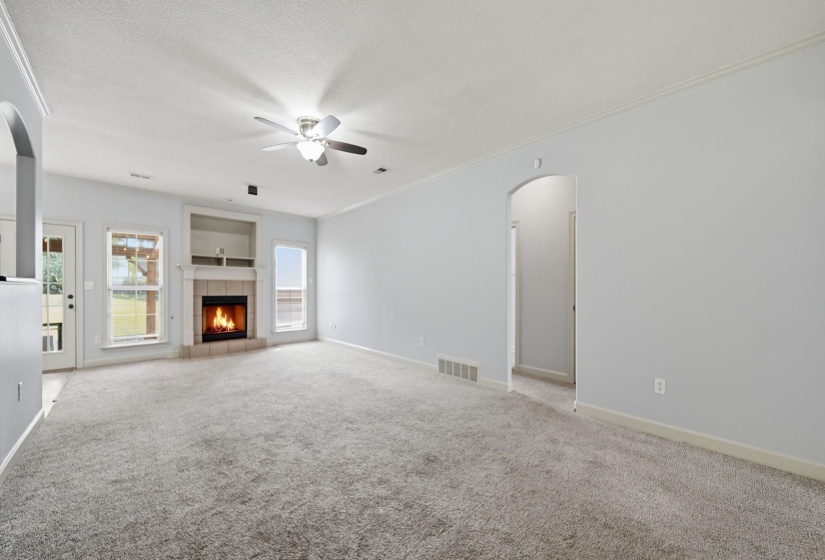 Spacious Great Room featuring light gray walls, plush gray carpeting, a built-in fireplace  & gas logs with a stone surround, an integrated ceiling fan with lighting, and multiple windows providing natural illumination