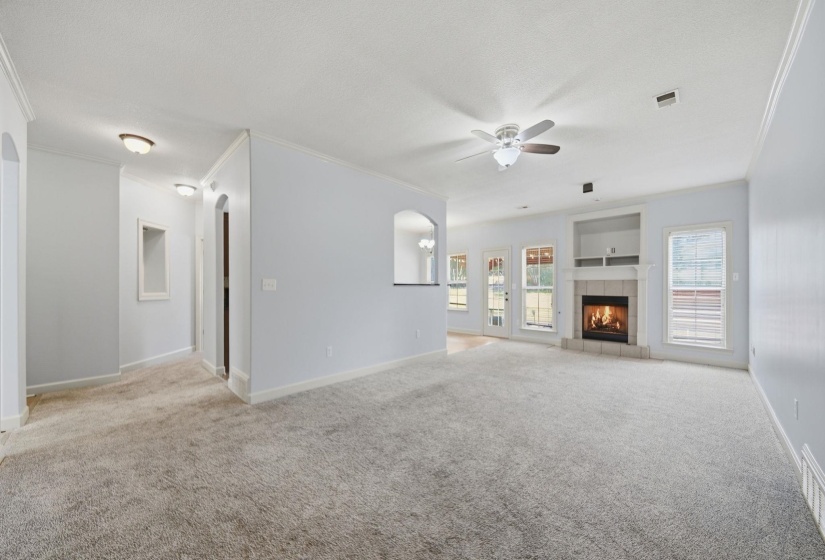 Spacious living area featuring light gray carpeting, a built-in fireplace with a mantel, multiple large windows, and a ceiling fan