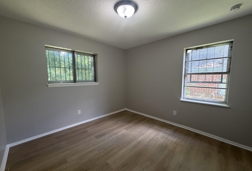 Room featuring wood-finish flooring, two windows with security bars, painted gray walls, white baseboards, and a flush-mount ceiling light
