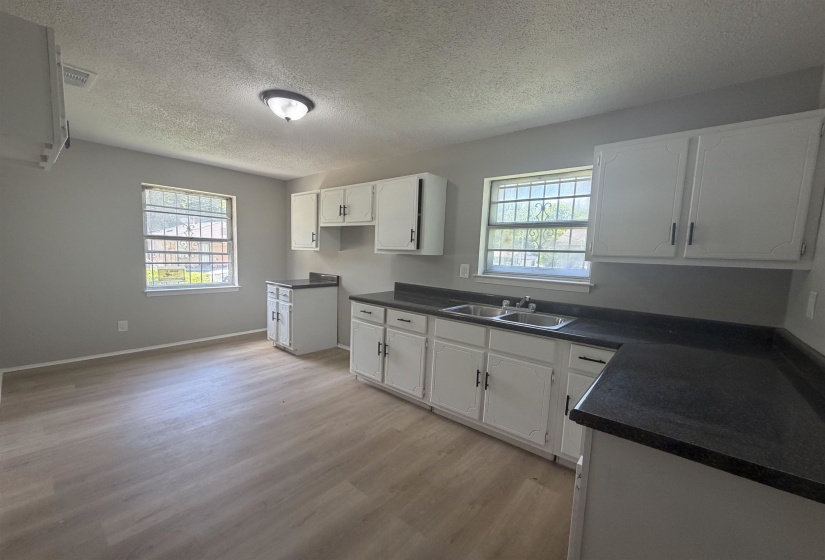 Bright kitchen featuring light wood-finish flooring, white cabinetry with dark hardware, dark countertops, and a dual basin stainless steel sink