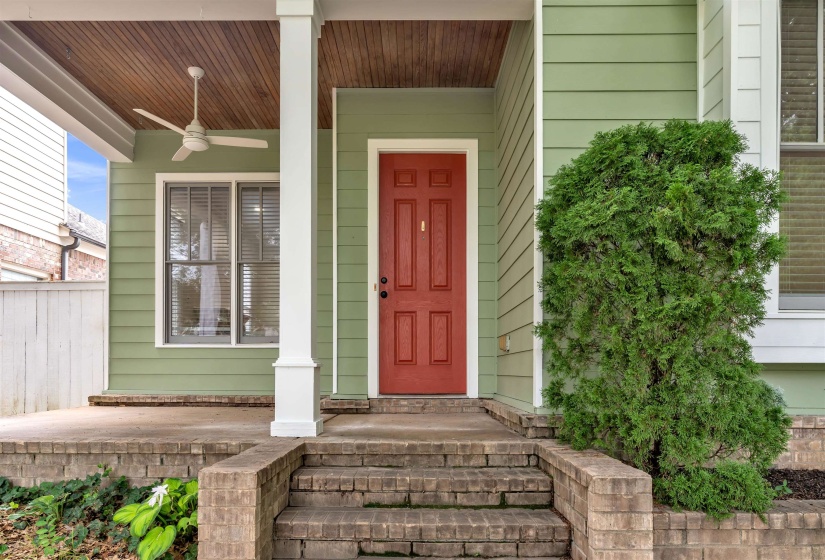 Covered front porch featuring a red entry door, light green siding, white columns, and a natural wood-finish ceiling with a ceiling fan