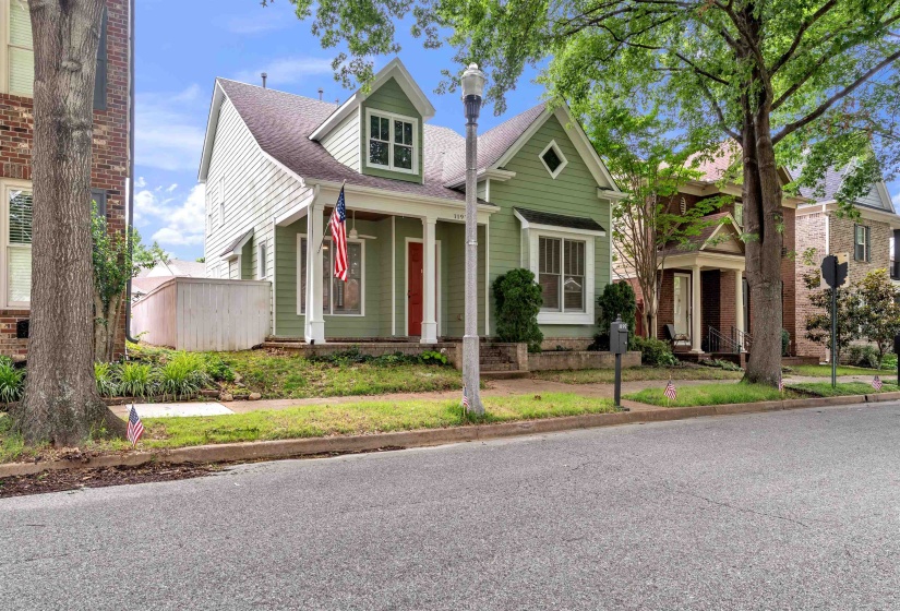 Green painted siding exterior featuring a covered front porch with white columns, a red entry door, a prominent front dormer, and a diamond-shaped window
