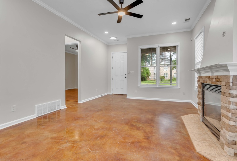 Living area featuring stained concrete flooring, a stone-faced fireplace with hearth, crown molding, and a ceiling fan with integrated lighting