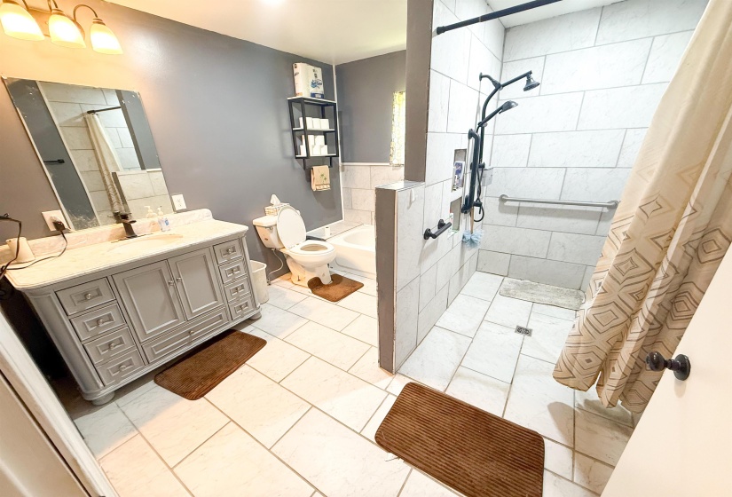 Bathroom featuring a light-toned vanity with a granite-style countertop, a multi-setting shower system, white tiled walls in the shower area, and light-colored floor tiling throughout