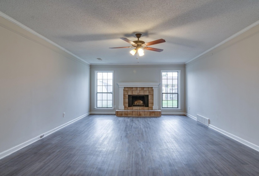 Spacious living area featuring neutral wall tones, wood-finish flooring, a brick fireplace with a white mantel, dual windows, and a ceiling fan with integrated lighting