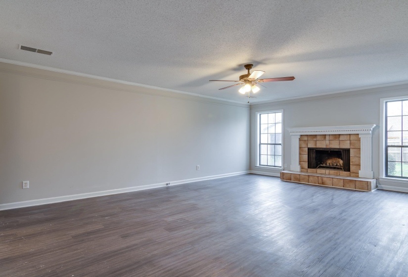 Spacious room featuring wood-finish flooring, a stone-tiled fireplace with a white mantel, multiple windows, a ceiling fan with light fixture, and crown molding