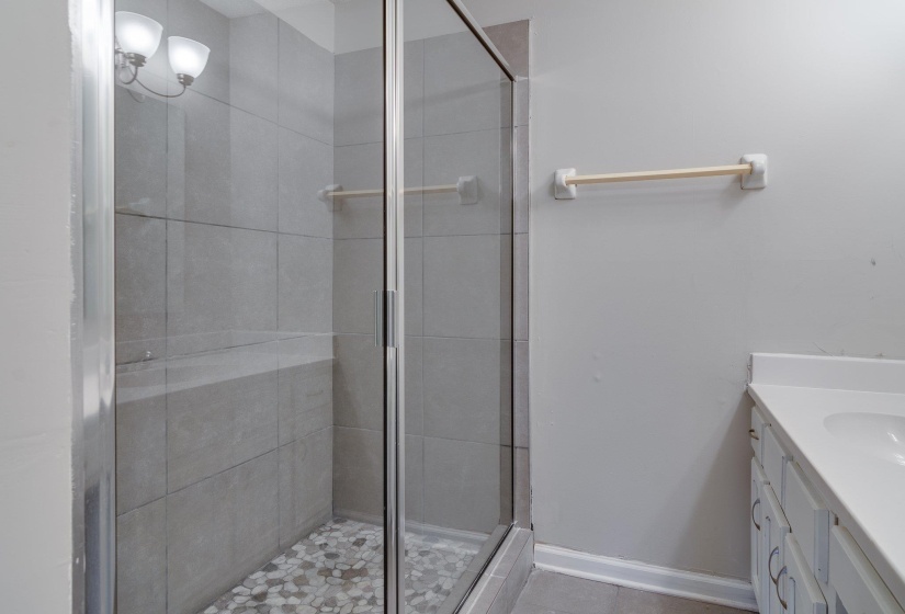 Glass-enclosed shower with large format grey tile surround, pebble tile flooring, and integrated shower bench