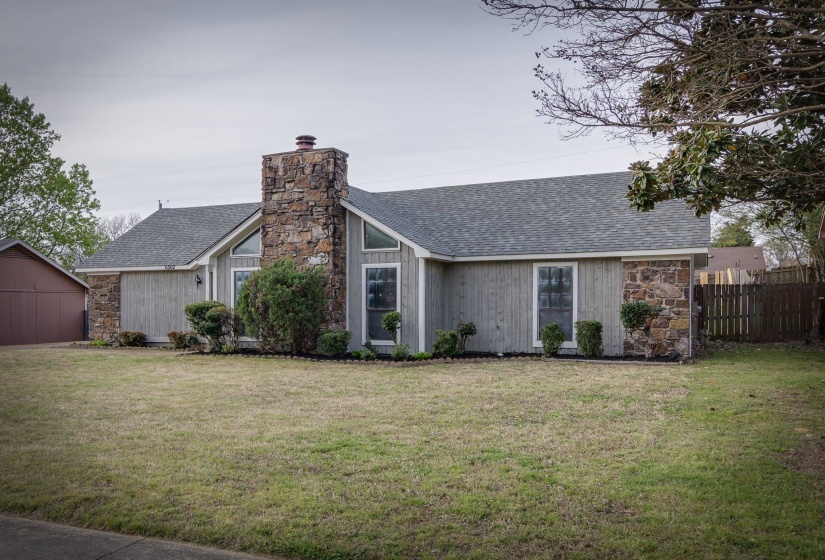 Exterior featuring a prominent stone chimney, vertical siding, a grey shingle roof, and a mature front yard