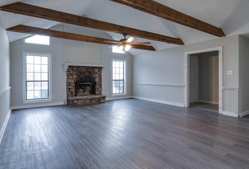 Spacious living area featuring a vaulted ceiling with exposed wood beams, a stone fireplace, wood-finish flooring, and multiple windows