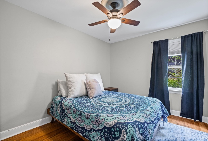 Bedroom with light gray walls, wood-finish flooring, a ceiling fan with integrated lighting, and a window featuring blinds and dark curtains
