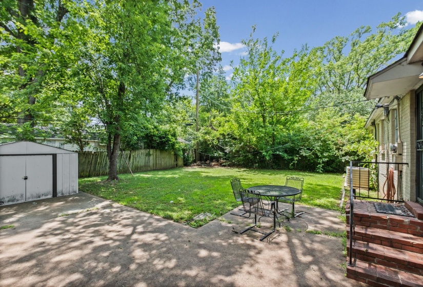 Expansive backyard featuring a concrete patio, a detached storage shed, lush green lawn, mature trees, and a wood privacy fence