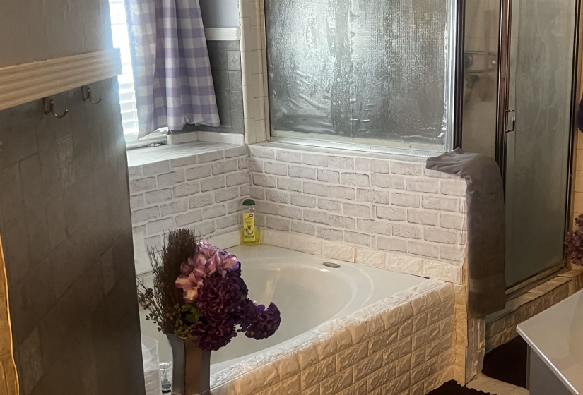 Built-in corner bathtub with brick-patterned surround, framed by a window with light-filtering blinds