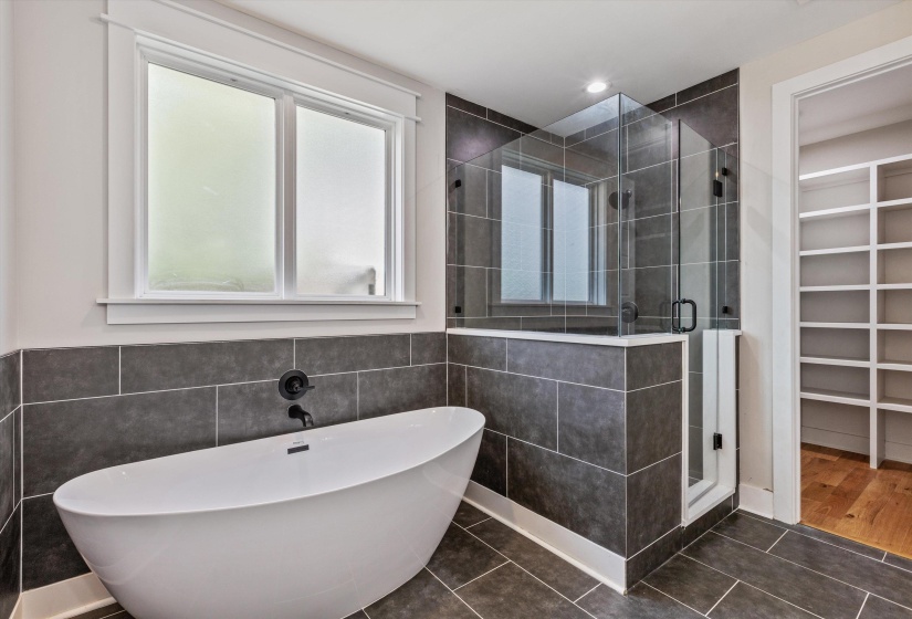 Contemporary bathroom featuring a freestanding soaking tub, a frameless glass shower enclosure, dark rectangular wall and floor tiling, a window with privacy glass, and a doorway leading to a room with built-in shelving and wood-finish flooring