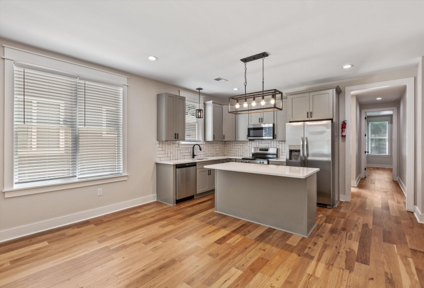 Open-concept kitchen featuring an island with a light-colored countertop, light gray cabinetry, stainless steel appliances, and a white herringbone tile backsplash