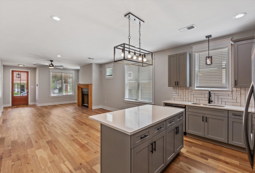 Open-concept living area with wood-finish flooring, a contemporary kitchen island with a light-colored countertop, and a fireplace with a wood surround