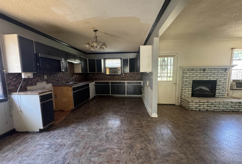 Open-concept living area featuring a brick-surround fireplace with hearth, a kitchen with black cabinetry and a built-in microwave, a ceiling-mounted light fixture, and wood-finish flooring throughout
