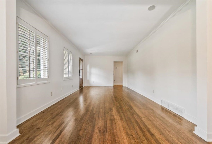 Expansive room featuring rich wood flooring, white walls, two windows with plantation shutters, a recessed ceiling speaker, and crisp crown molding