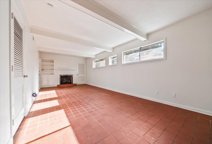 Spacious interior featuring terra cotta tile flooring, exposed ceiling beams, a built-in fireplace, integrated shelving, and multiple horizontal windows
