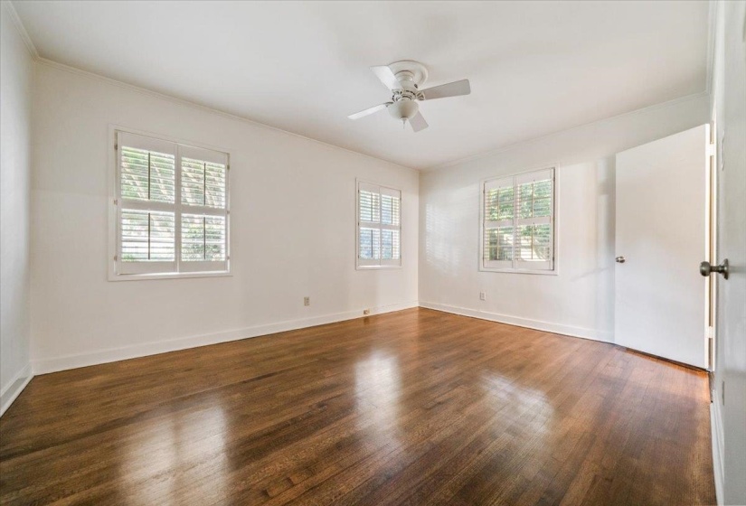 Hardwood flooring throughout, featuring multiple windows with plantation shutters, a ceiling fan, and white baseboards