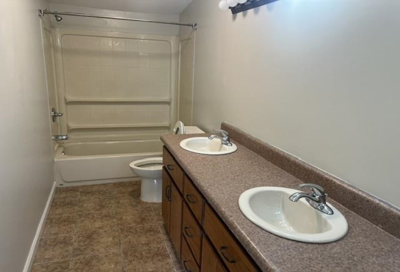 Bathroom featuring a dual vanity with laminate countertops, two undermount sinks with chrome fixtures, wood-finish cabinetry, and a full tub/shower combination with a textured surround
