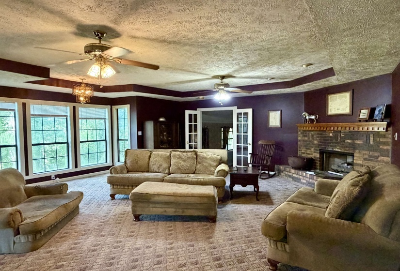 Spacious living area featuring a prominent brick fireplace with a wood mantle, multiple ceiling fans, and recessed tray ceiling details