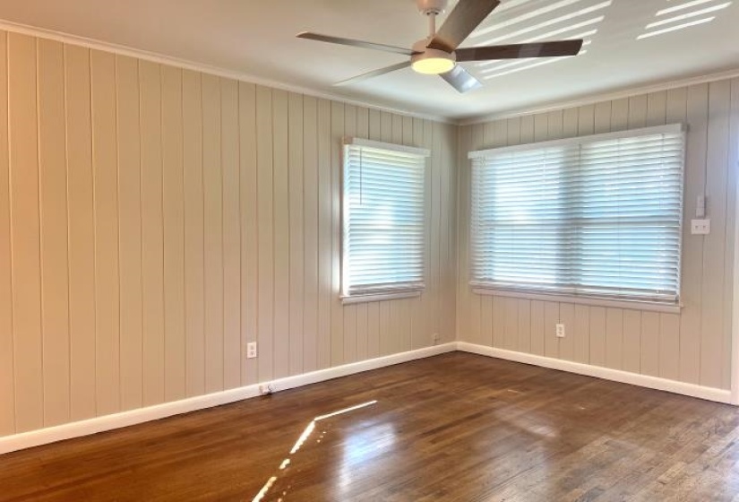 Room with wood-finish flooring, two windows with blinds, a ceiling fan with integrated lighting, and vertical paneling on the walls