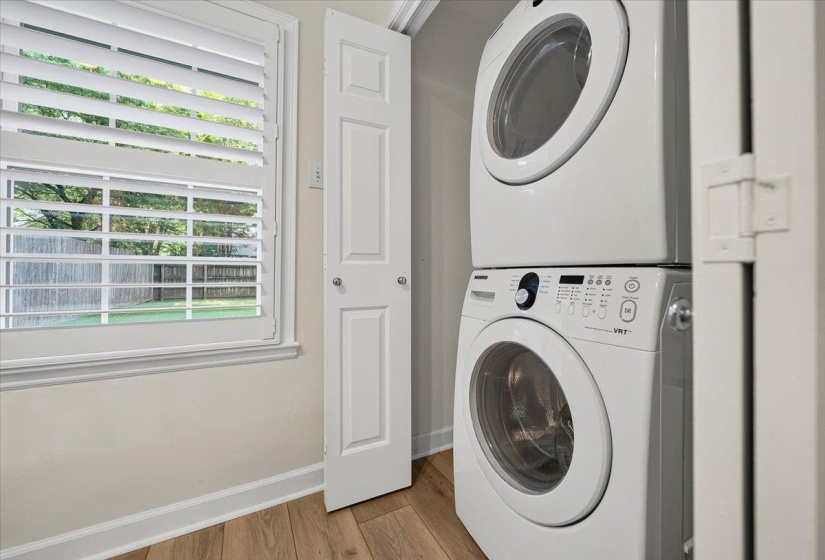 Dedicated laundry area featuring a stacked washer and dryer, wood-finish flooring, white plantation shutters, and a paneled bifold door