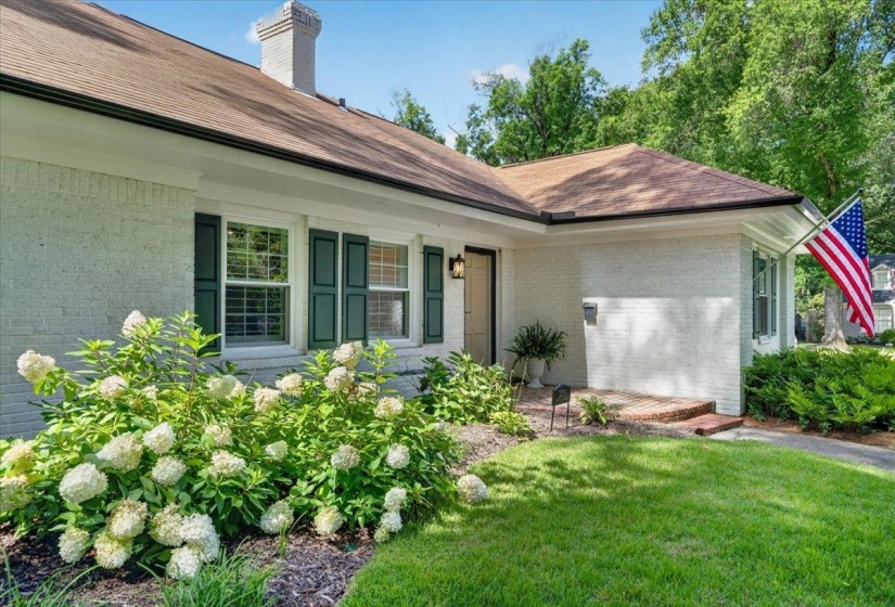White painted brick exterior featuring dark green shutters, a brown shingle roof, and a brick entrance path