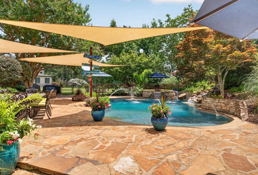 Outdoor entertaining space featuring a freeform pool with stone waterfall, flagstone patio, multiple shade sails, and mature landscaping