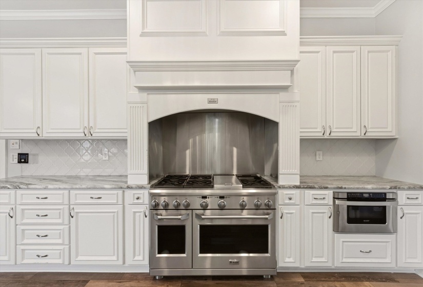 Gourmet kitchen featuring a stainless steel professional range with a custom decorative hood, built-in microwave, white shaker cabinetry, patterned tile backsplash, and stone countertops