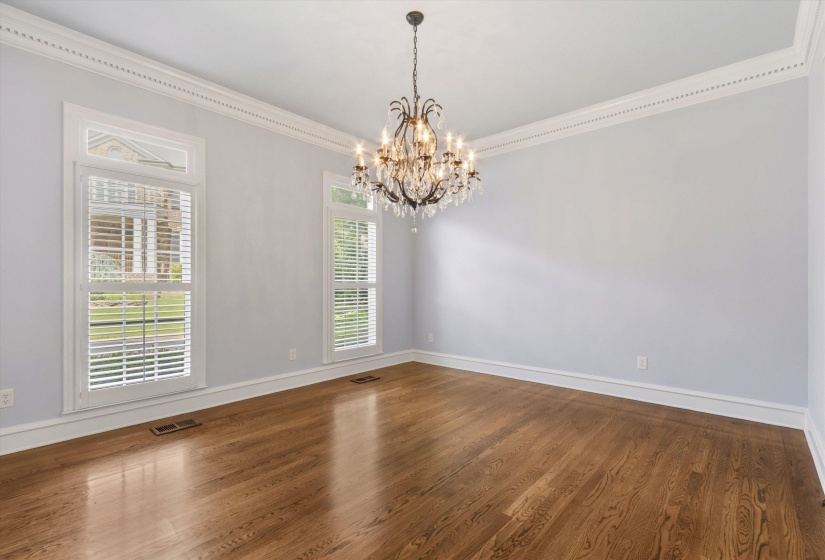 Spacious room featuring wood-finish flooring, a tiered crystal chandelier, and two windows with white plantation shutters