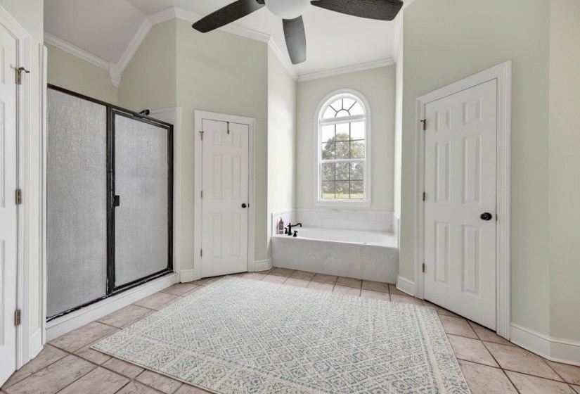 Spacious room featuring a built-in tub with an arched window, a separate shower with opaque glass, tile flooring, and a ceiling fan