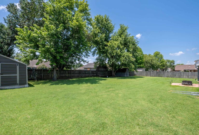 Spacious backyard featuring a large green lawn, mature trees providing natural shading, a privacy fence, and a metal storage shed