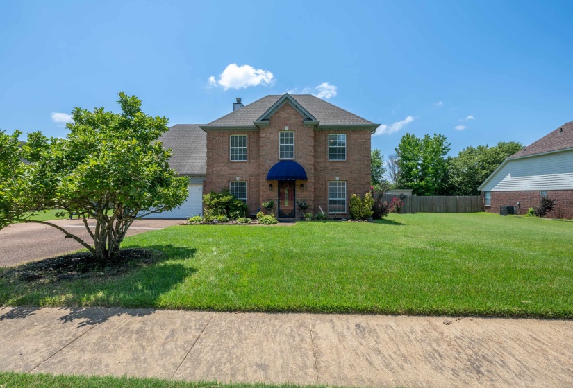 Traditional brick exterior featuring a blue entrance awning, a two-car garage, mature landscaping, and an expansive front lawn
