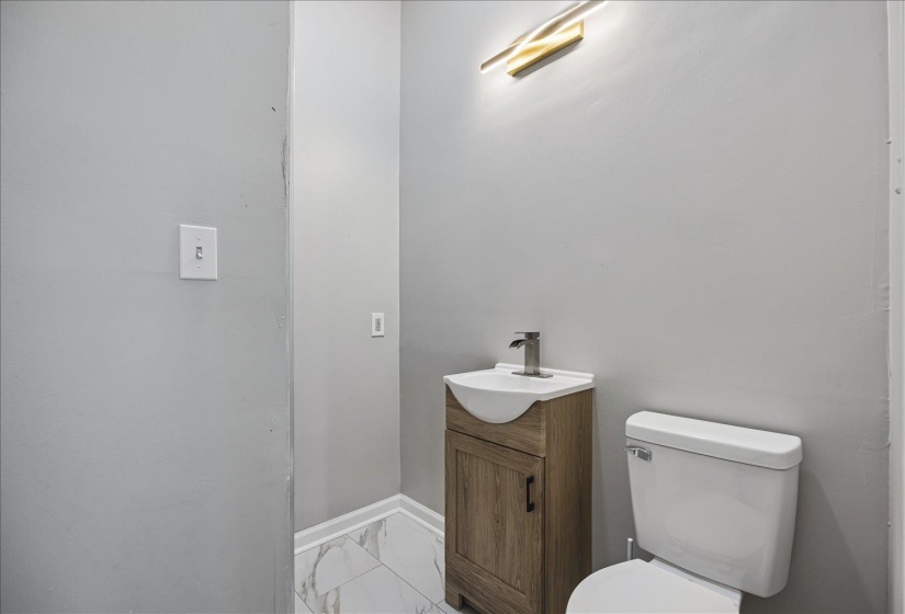 Powder room featuring a wood-finish vanity with an integrated white sink, a modern gold-toned wall sconce, and large format floor tiling