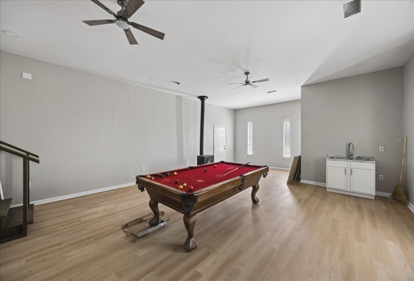 Spacious bonus room featuring wood-finish flooring, two ceiling fans, a functional wet bar with a sink and white cabinetry, and a visible staircase