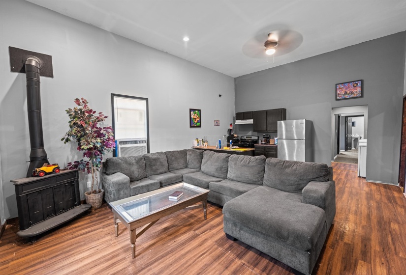 Open concept living area featuring wood-finish flooring, a freestanding stove with a black pipe, and a kitchen area with dark cabinetry, stainless steel refrigerator, and a built-in overhead microwave