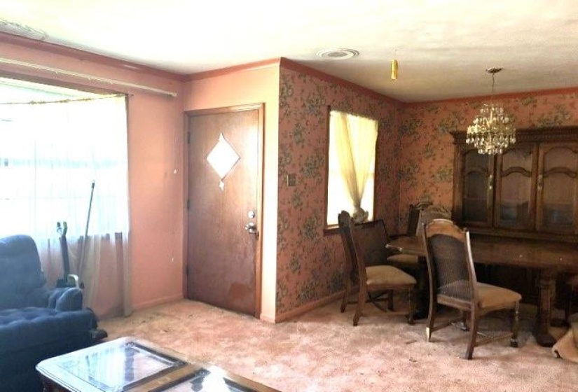 Combined living and dining area featuring a front entry door with decorative glass insert, a chandelier lighting fixture, and a window with sheer curtains
