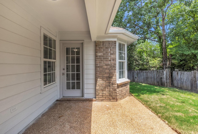 Covered patio featuring an exposed aggregate concrete surface, leading to a bay window with brick facade and a wood privacy fence