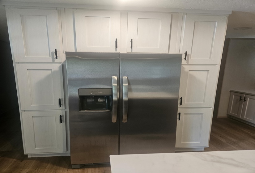 Stainless steel side-by-side refrigerator with exterior ice and water dispenser, flanked by white shaker-style cabinetry with black hardware, wood-finish flooring, and a white countertop