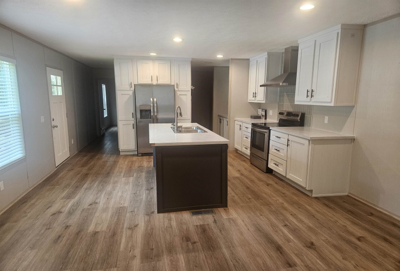 Spacious kitchen featuring an island with an integrated sink, stainless steel appliances, white cabinetry, light-toned countertops, and wood-finish flooring