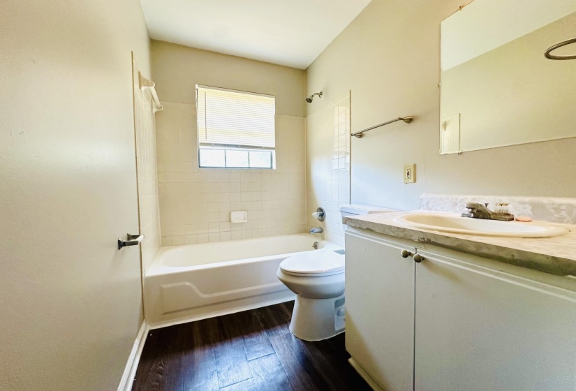 Bathroom featuring a single vanity with a white sink, an integrated bathtub and shower, a window with blinds, tiled shower surround, and wood-finish flooring