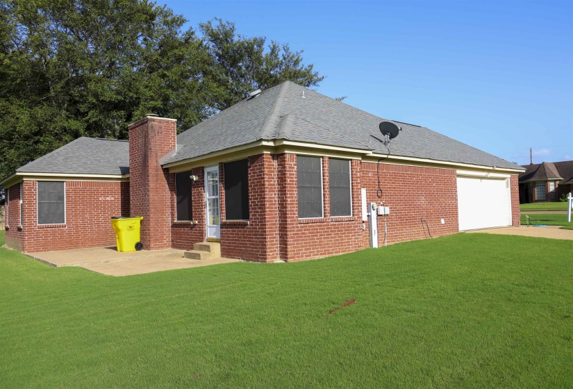 Brick exterior with a gray shingled roof, featuring a chimney, a concrete patio, a single-car garage, and a lush green lawn