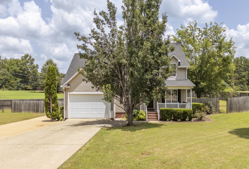 Two-story residence featuring light-colored siding, a covered front porch with white railings, a front-facing attached garage, a paved driveway, and mature landscaping