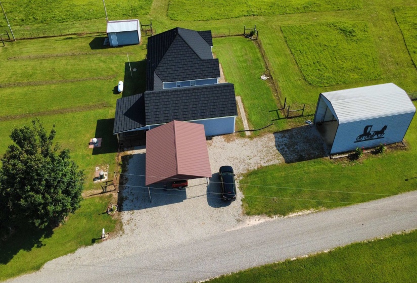 Blue exterior residence featuring a dark shingled roof, an attached carport with a red metal roof, a gravel driveway, and a detached metal storage building