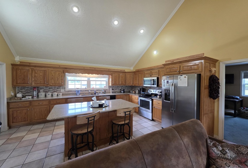 Kitchen featuring vaulted ceiling with recessed lighting, wood cabinetry, stainless steel appliances, a multi-tone tile backsplash, and a central island with countertop seating